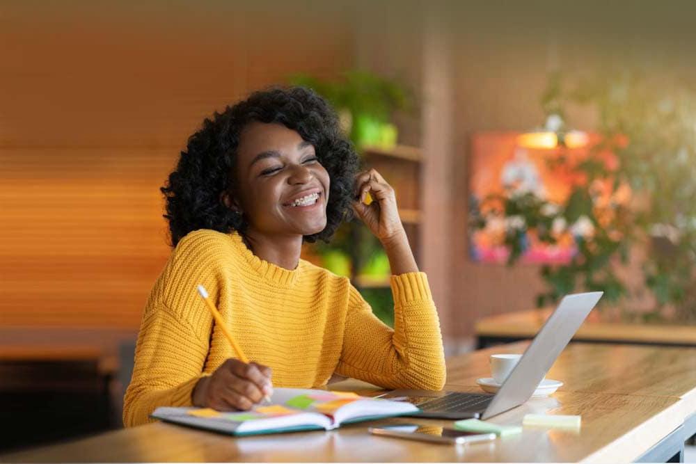 Young girl smiling while taking note in a notebook while looking at a computer wearing a yellow sweater taking an online class at Ohio's Great River Connections Academy